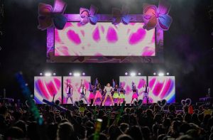 Jojo Siwa (center) and her dancers, including Loy Norrix alumna Taylor Timmerman, stand with an arm raised high in front of the crowd at a show on the D.R.E.A.M. tour.  The stage is decorated with bows, hearts and pink screens.  