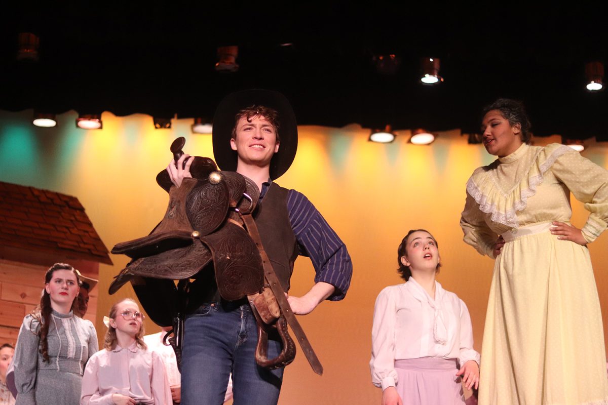 Braeden Davis holds up a horse saddle in his role of Curly.  Members of the cast look at him inquisitively, as Hana Westrick, playing Aunt Eller, speaks to him.  
