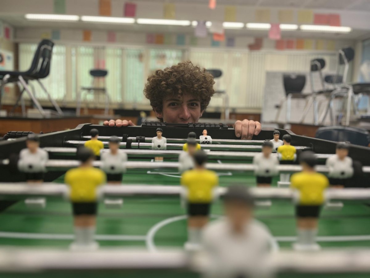 Photo Illustration. Foosball club leader, sophomore Felix Bahrou, peers over the edge of the foosball table in B-14. This year, the foosball club met on Mondays, immediately after school.